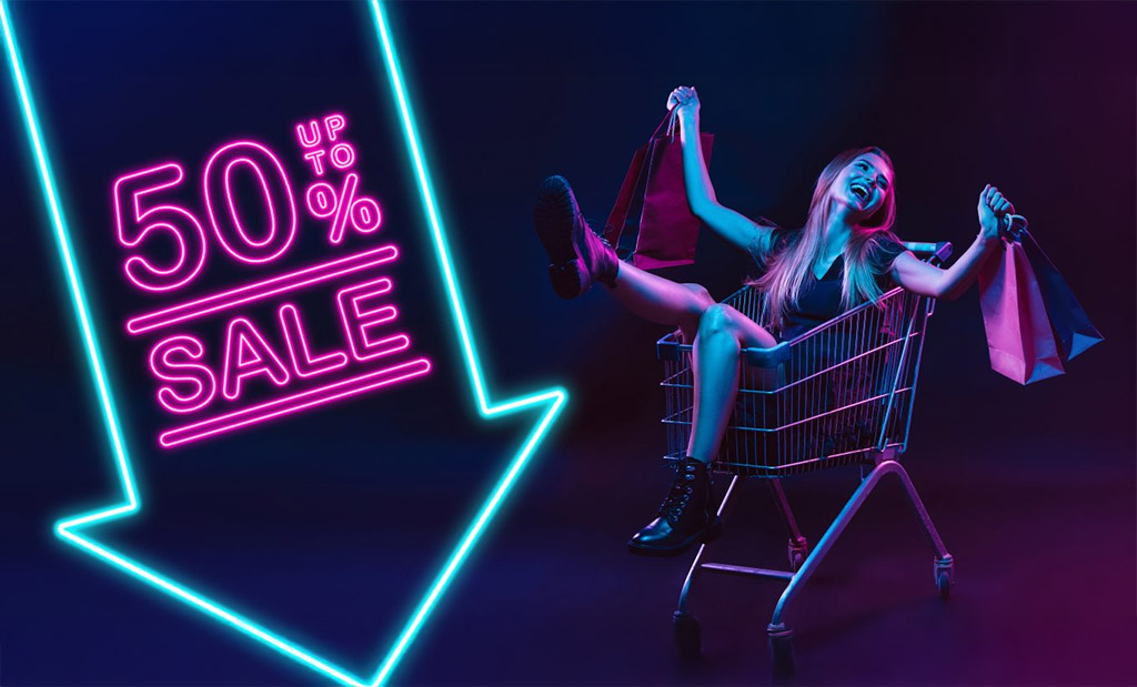 Sötét háttér, bal oldalon nagy lefele mutató kék neon nyíl, benne rózsaszín neon felirat. up to 50% sale. Jobb oldalon egy bevásárló kosárban ül egy fiatal nevető hölgy. Kezében bevásárló táskák. 