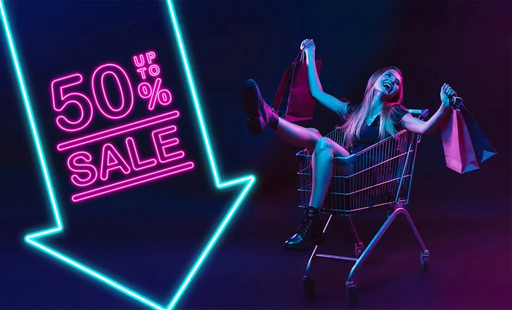 Sötét háttér, bal oldalon nagy lefele mutató kék neon nyíl, benne rózsaszín neon felirat. up to 50% sale. Jobb oldalon egy bevásárló kosárban ül egy fiatal nevető hölgy. Kezében bevásárló táskák. 