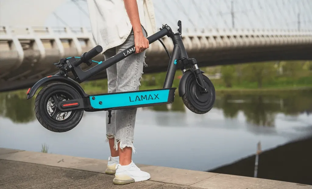 Egy személy látható, amint egy összecsukott, fekete-türkiz színű Lamax eCruiser SC30 elektromos rollert visz a kezében egy vízparti sétányon, a háttérben egy híd sziluettjével.