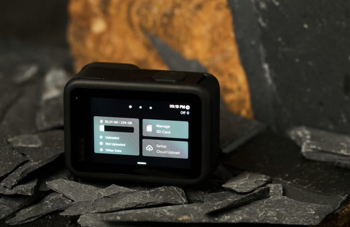 GoPro HERO13 Black akciókamera hátulról, a bekapcsolt érintőképernyőn a memóriakártya és a felhő alapú feltöltés beállításai látszanak sötét palaköveken elhelyezve.