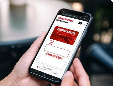Kézben tartott okostelefonon a Media Markt ajándékkártya oldala látható, vörös és fehér design elemekkel.