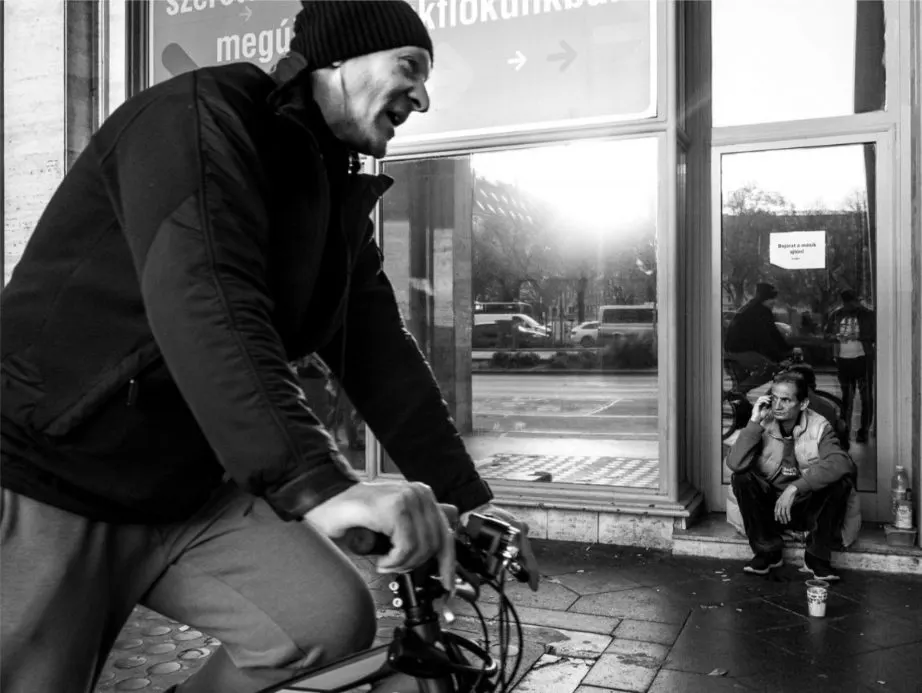 Fekete-fehér utcai pillanatkép: egy kerékpáros halad el az előtérben, miközben a háttérben egy kirakat mellett egy férfi ül a földön és telefonál.