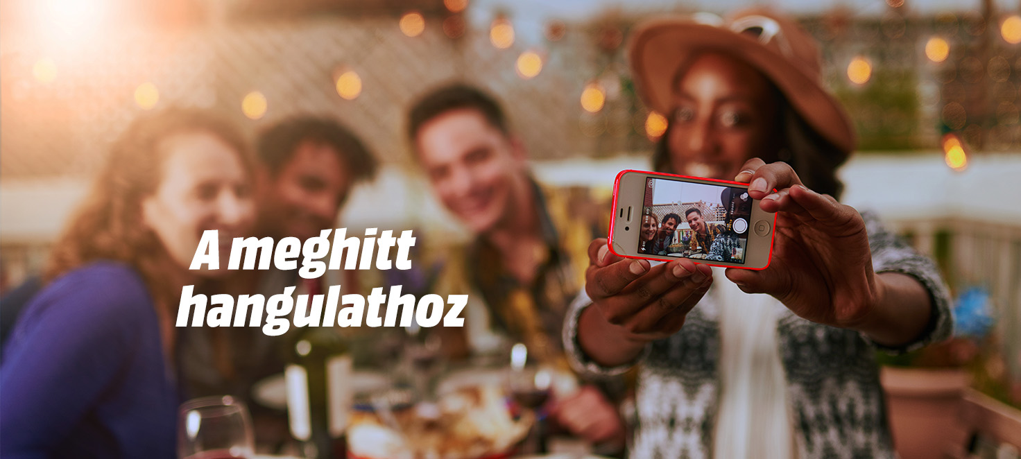 Meghitt hangulathoz feliat, csoport emberek ül az asztalnál, és egy nő fényképet készít egy telefonnal. Fényfüzérek vannak a háttérben.