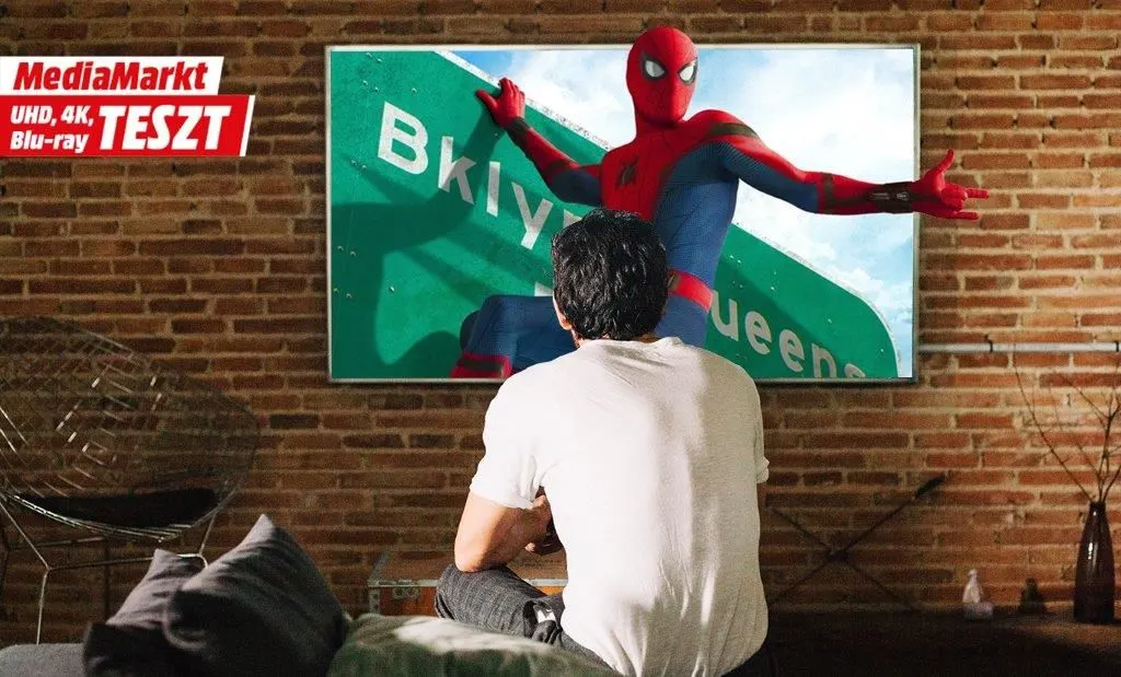 Egy ember háttal ülve nézi a TV-t, a képernyőn Pókemberrel. A TV felett a MediaMarkt UHD, 4K, Blu-ray TESZT felirat látható.