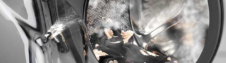 Ezüstös mosógép, benne fekete ruha, sárga mintákkal, vízpermettel.