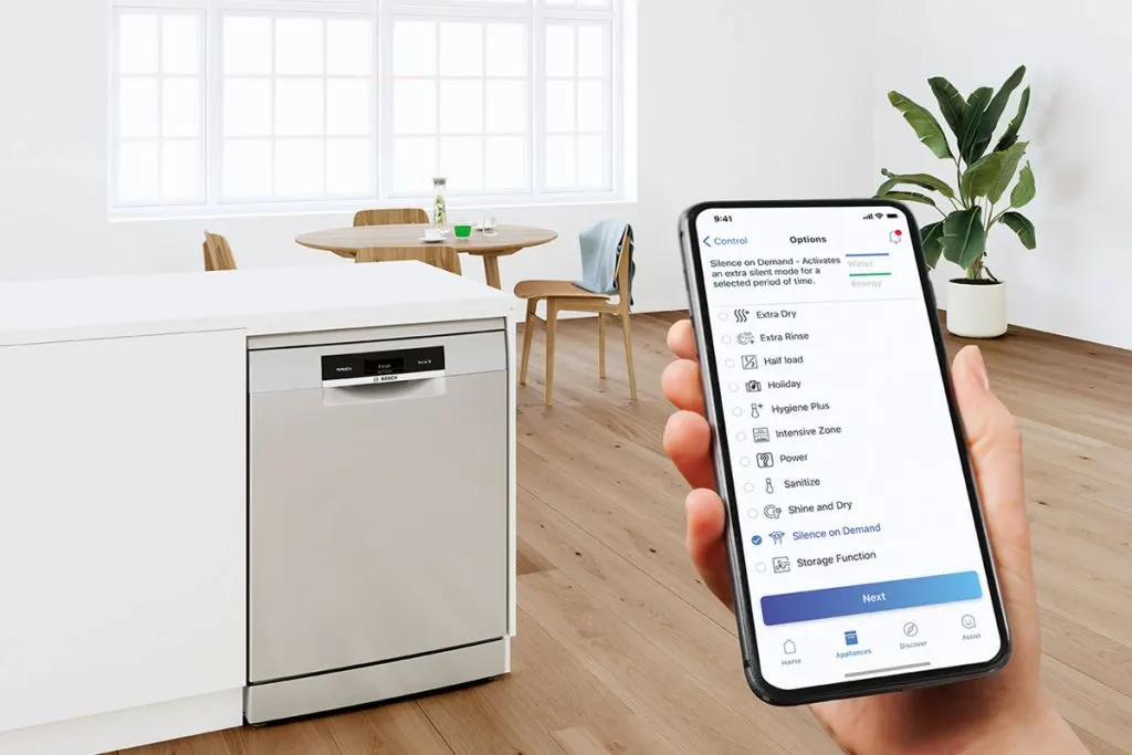 Nő használja az okostelefonos Home Connect alkalmazást egy intelligens mosogatógép vezérlésére a konyhában, ahogy a milyen-az-intelligens-mosogatogep-home-connect-1024x683.jpg fájlban látható.
