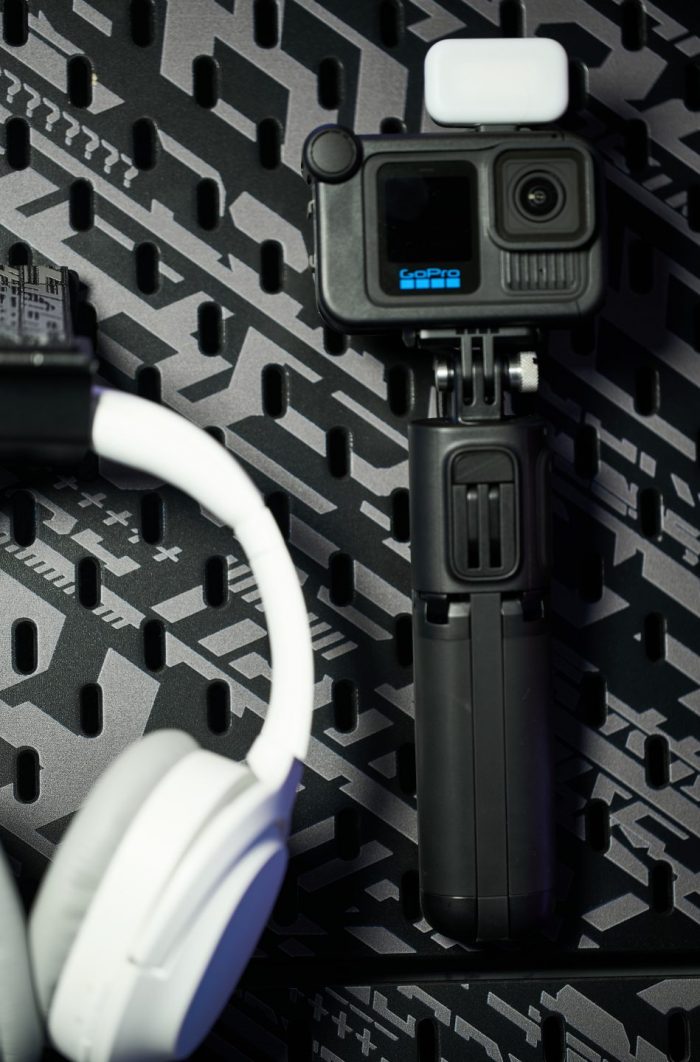 GoPro HERO13 Black akciókamera Media Mod kiegészítővel és egy kis méretű LED lámpával felszerelve, egy összecsukható tripod markolaton, mellette egy fehér fejhallgatóval szürke mintás háttér előtt.