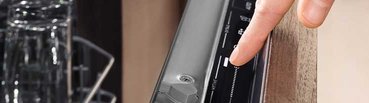 Ujj érinti a mosogatógép fekete vezérlőpaneljét, mely ezüst szegélyű. Üvegek láthatók a háttérben.