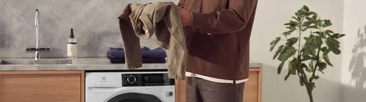 Egy személy ruhát tart mosógép előtt. A háttérben mosogató, növény és szekrény látható.