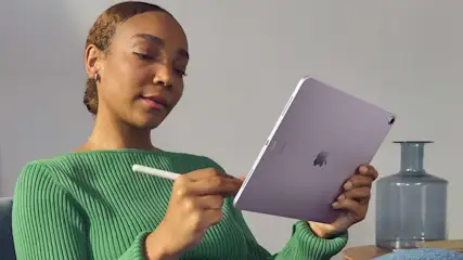 Frau mit grünem Pullover hält ein lila Tablet und einen weißen Stift. Vase im Hintergrund.