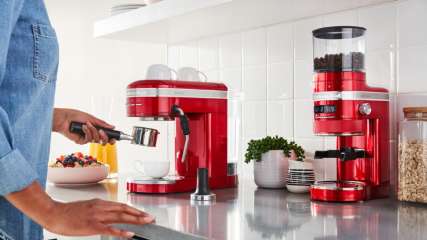 Frau bedient bei ihr zuhause eine rote Siebträgermaschine der Marke KitchenAid. Auf der Küchenarbeitsfläche ein Gerät zum Mahlen von Kaffeebohnen, ebenfalls von KitchenAid. steht außerdem 