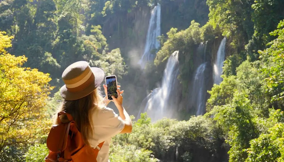 Eine Person mit Hut und Rucksack fotografiert einen Wasserfall mit einem Mobiltelefon. Bäume und grüne Vegetation umgeben die Szene.