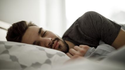 Junger Mann liegt im Bett mit geschlossenen Augen und einem zufriedenen Gesichtsausdruck. 