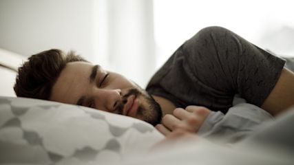 Junger Mann liegt im Bett mit geschlossenen Augen und einem zufriedenen Gesichtsausdruck.