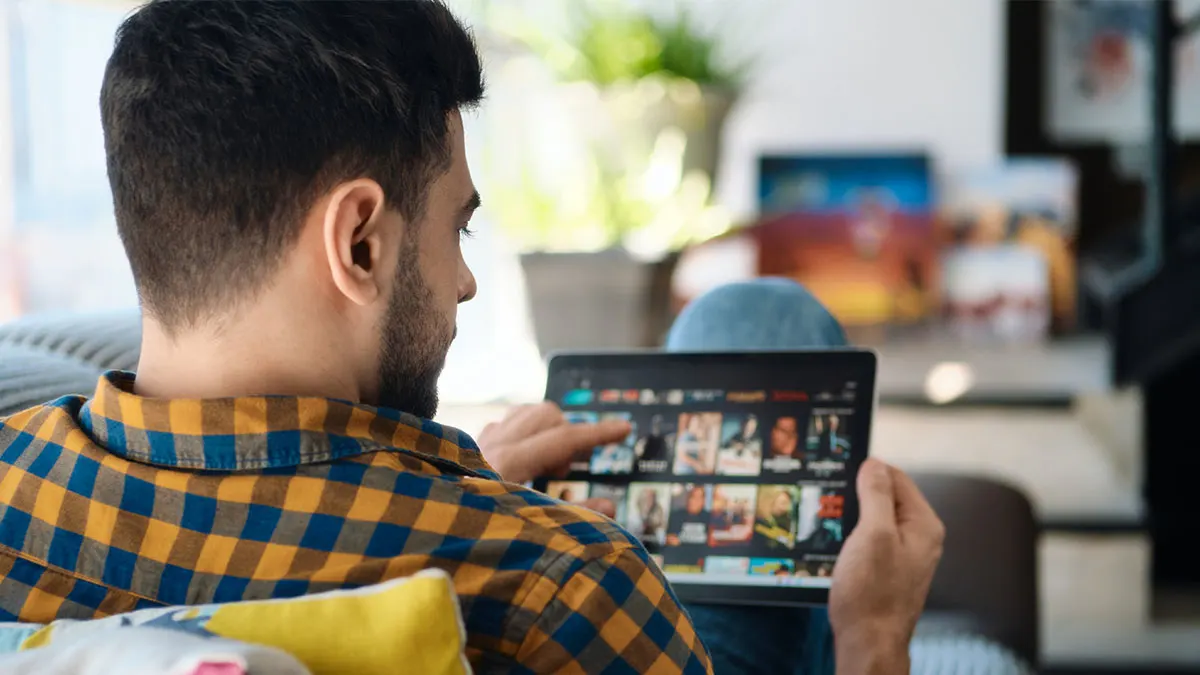 Ein Mann sitzt auf einem Sofa und wählt auf dem Tablet einen Film oder eine Serie in einem Streaming-Dienst aus.