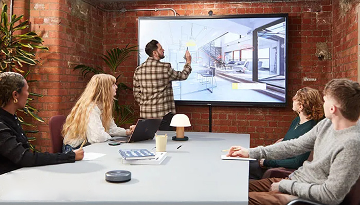 Eine Gruppe von Personen sitzt an einem Tisch vor einem großen Bildschirm, auf dem ein Gebäudeentwurf angezeigt wird.