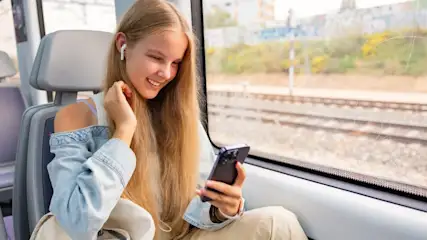 Ein Mädchen mit Kopfhörern sitzt in einem Zug, blickt auf ein Smartphone und lächelt. Bahngleise sind durch das Fenster sichtbar.