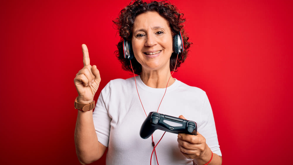 Eine Frau im mittleren Alter lächelt mit erhobenem Zeigefinger und Spielecontroller