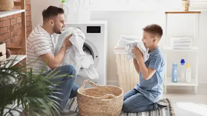 Vater lädt Wäsche in Waschmaschine, während Sohn mit Handtuch spielt, Korb und Regal im Raum.