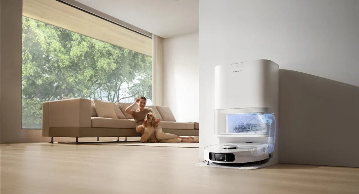 Der Saug-Wisch-Roboter Dreame L10s Ultra Gen2 steht in seiner Ladestation, im Hintergrund sitzen eine Frau und ein Hund vor einem Sofa.