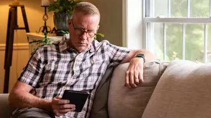 Ein Mann mit kariertem Hemd sitzt auf einem Sofa und liest auf einem schwarzen Tablet. Im Hintergrund steht eine Lampe und ein Fenster.