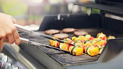 Eine Grillplatte mit Burgern und Gemüsespießen wird mit einer Zange von einer Hand bewegt.