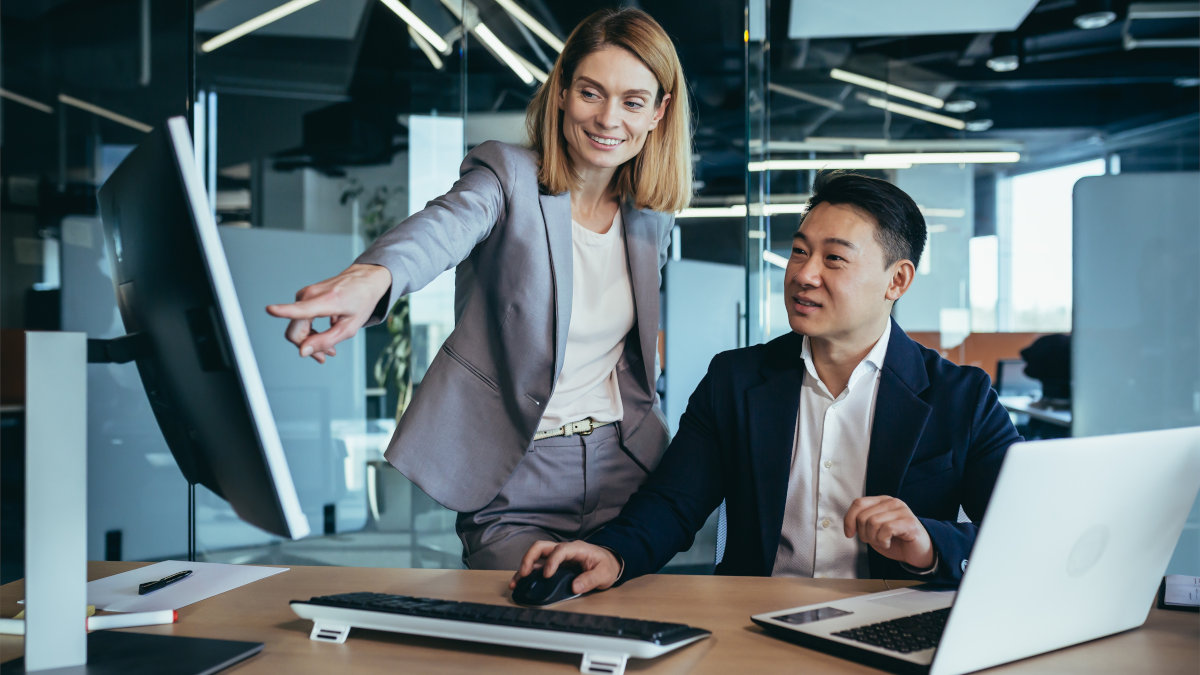 Eine stehende Frau im Anzug zeigt auf einen Monitor, während ein Mann im Anzug neben ihr an einem Schreibtisch mit Laptop sitzt.