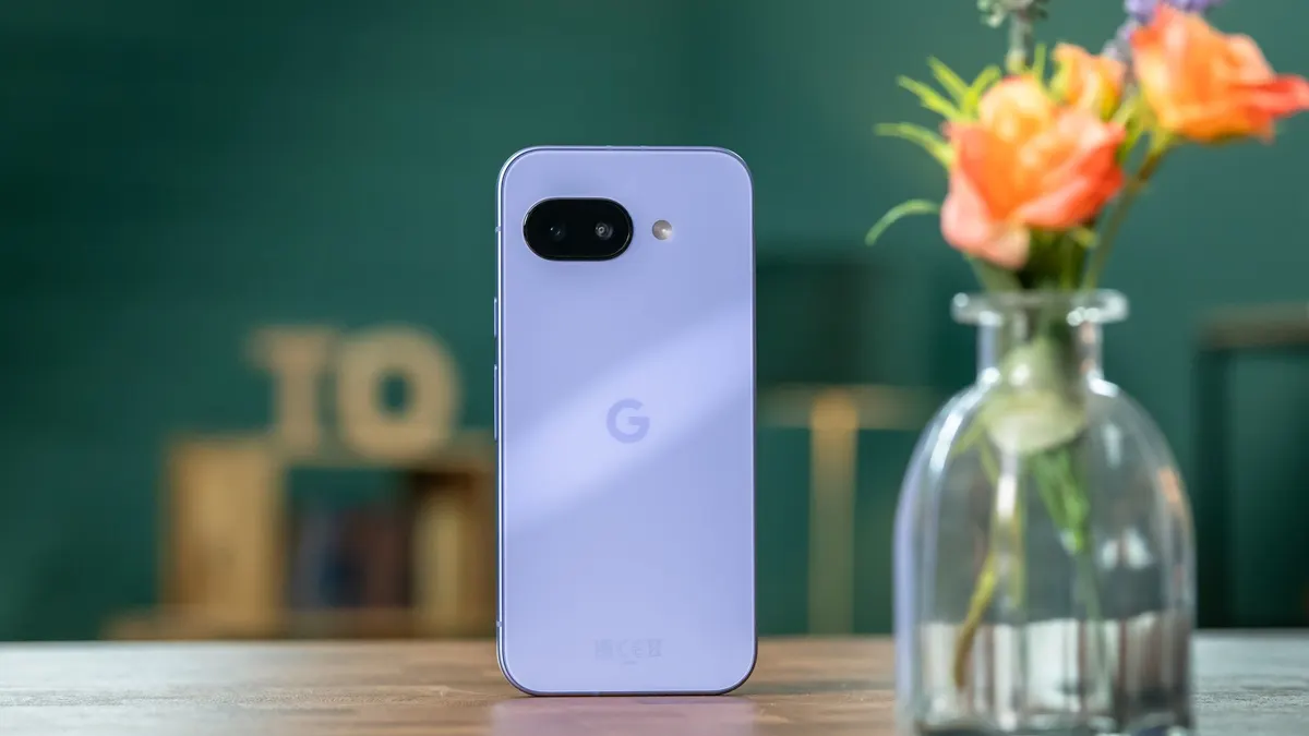 Ein lila Smartphone mit Google-Logo steht neben einer Glasvase mit orangefarbenen Blumen auf einem Holztisch vor einem grünen Hintergrund.
