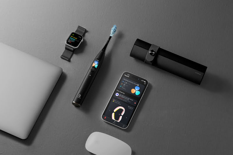 Auf grauem Grund liegen Laptop, Uhr, Zahnbürste, Hülle, Handy und Maus. Auf den Bildschirmen sind Daten zu sehen.