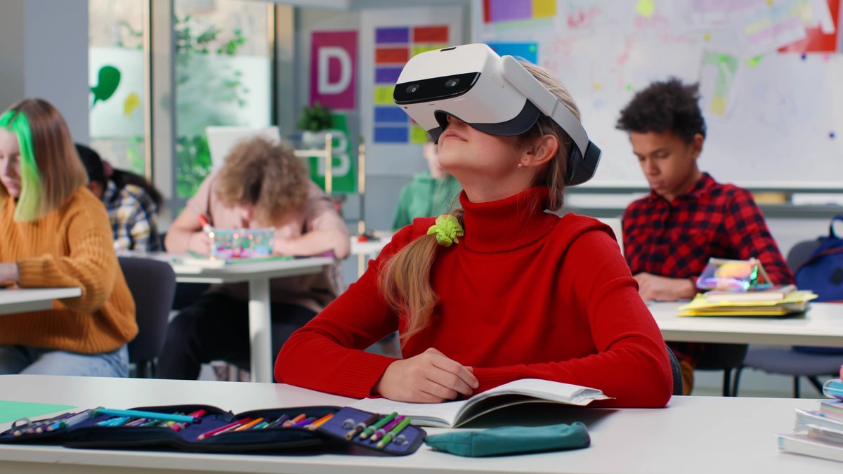 In einem Klassenzimmer sitzt ein Mädchen mit einem roten Rollkragenpullover und einem VR-Headset. 