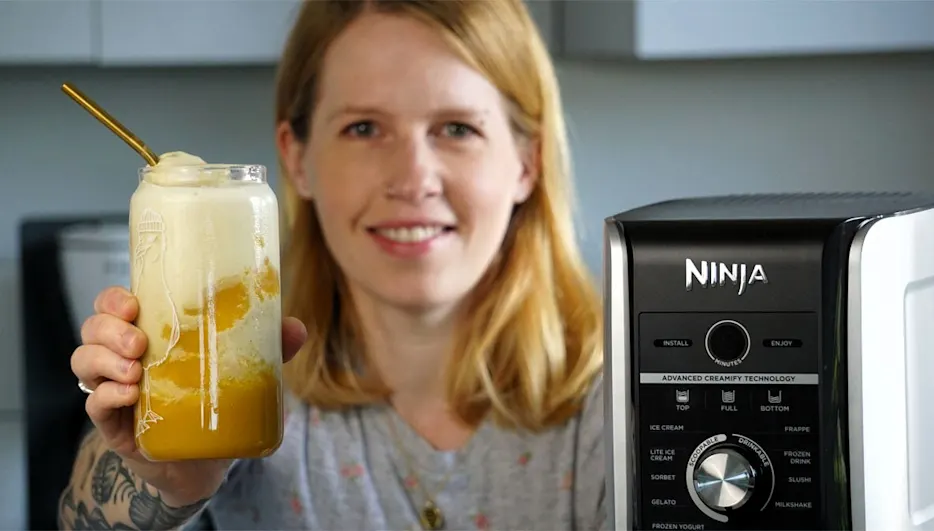 Franzi hält ein Glas mit einem Eisgetränk in der Hand, neben ihr steht die Ninja Creami Deluxe.