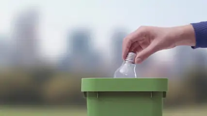 Eine Hand wirft eine Glühbirne in einen grünen Recyclingbehälter. Im Hintergrund sind verschwommene Gebäude zu sehen.