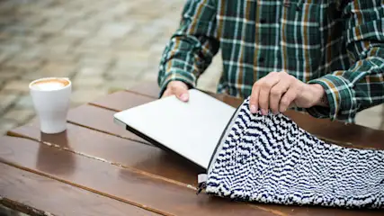 Eine Person legt einen Laptop in eine blau-weiße Tasche auf einem Holztisch. Eine weiße Tasse steht daneben.