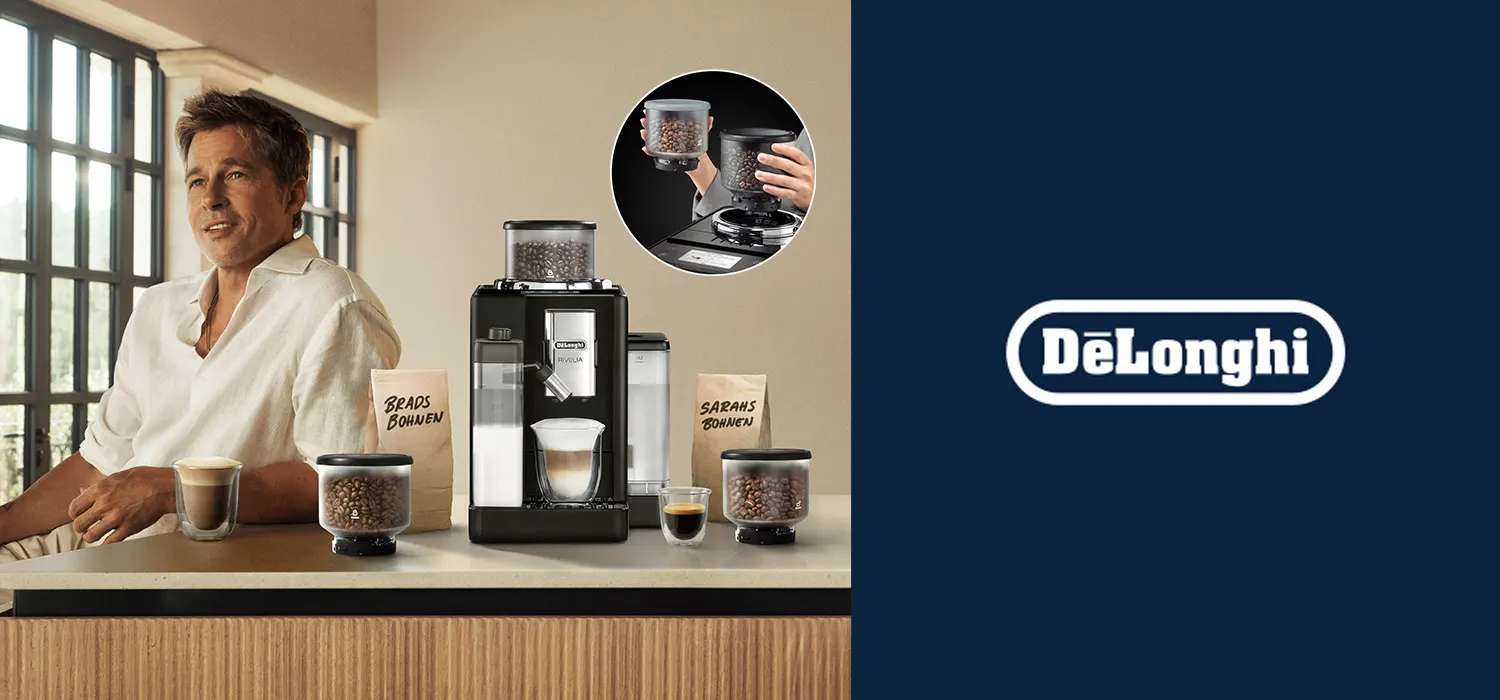 Eine Kaffeemaschine steht zwischen zwei Kaffeebohnenbehältern und zwei Tassen Kaffee auf einer Theke. Ein Mann sitzt daneben.