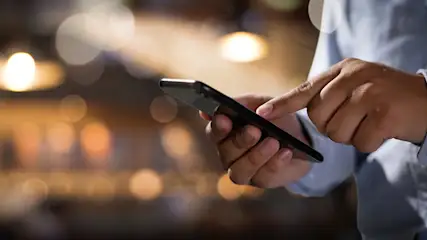 Eine Hand in hellblauem Hemd tippt auf ein schwarzes Smartphone. Im Hintergrund sind unscharfe Lichter.