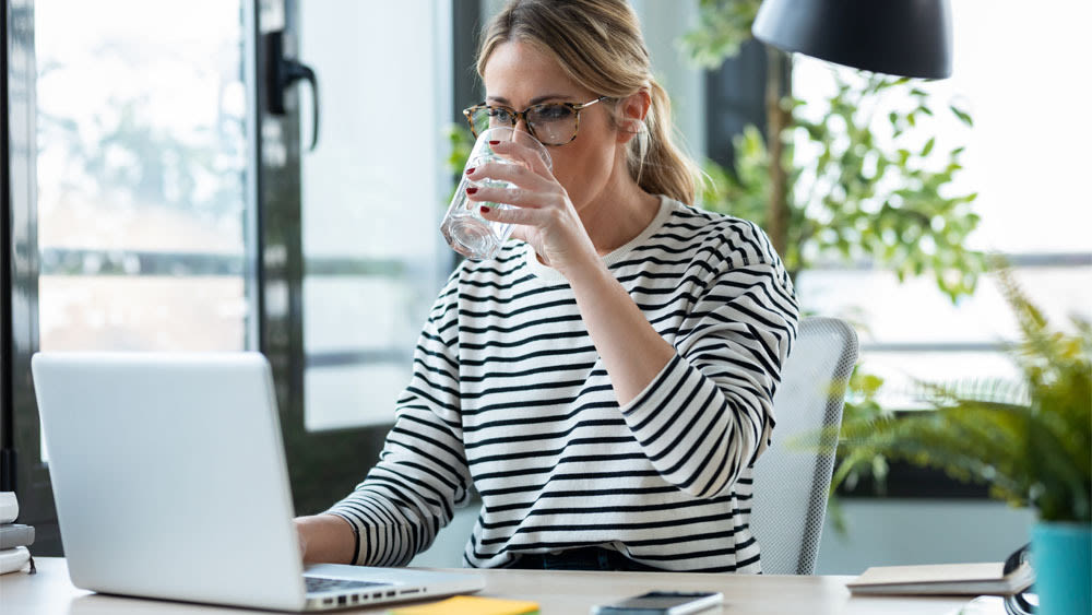 Eine Frau sitzt am Schreibtisch und trinkt Wasser aus einem Glas.