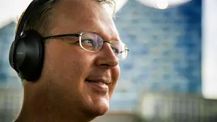Ein Mann mit Brille und Kopfhörern lächelt. Im Hintergrund ist ein verschwommener blauer Farbverlauf zu sehen.
