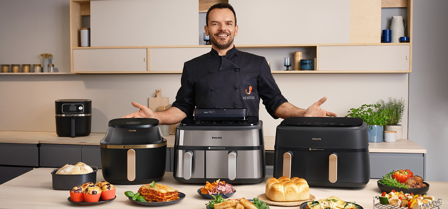Koch Steffen Henssler präsentiert drei Philips Airfryer auf einer Küchentheke. Im Vordergrund stehen verschiedene Speisen, die mit den Airfryern zubereitet wurden.