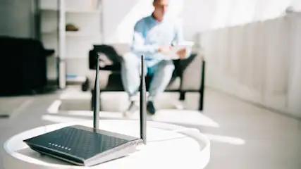 Ein schwarzer Router mit zwei Antennen steht auf einem weißen Tisch. Im Hintergrund sitzt eine Person auf einem Sofa.