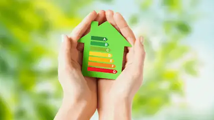 Zwei Hände halten ein grünes Hausmodell mit einer farbigen Energieeffizienzskala von A bis G vor verschwommenem grünem Hintergrund.