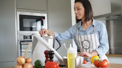 Eine Frau mit Schürze kocht mit einem Küchengerät. Zwiebeln und Gemüse liegen auf der Arbeitsfläche.