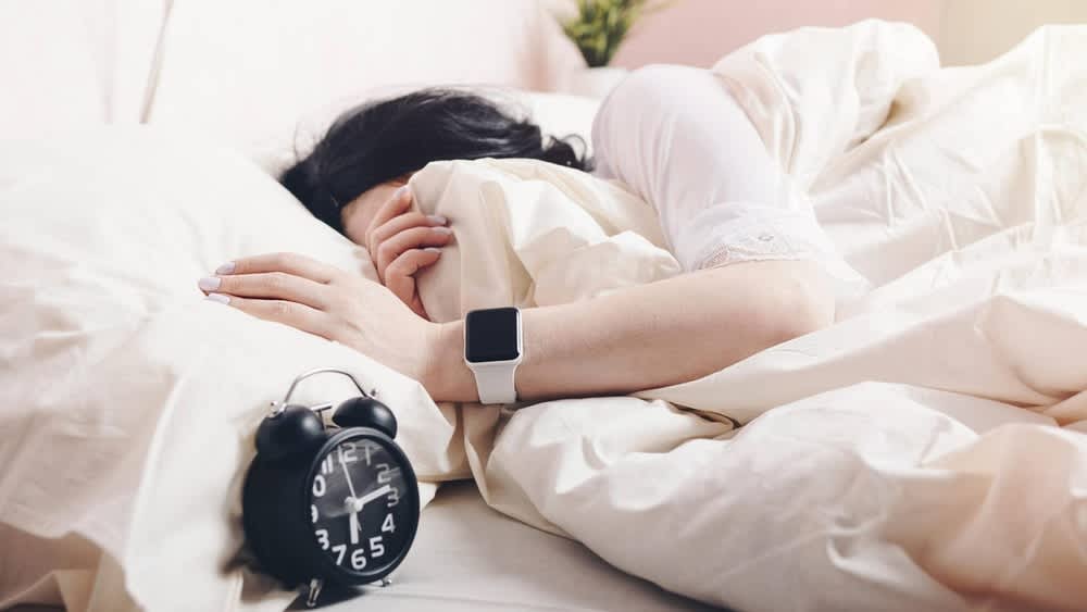 Frau liegt mit Smartwatch im Bett neben einem Wecker
