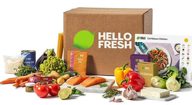 Ein Karton mit "Hello Fresh"-Logo umgeben von Zutaten wie Gemüse, Nudeln, Fisch und Gewürzen. Kochbox-Konzept.