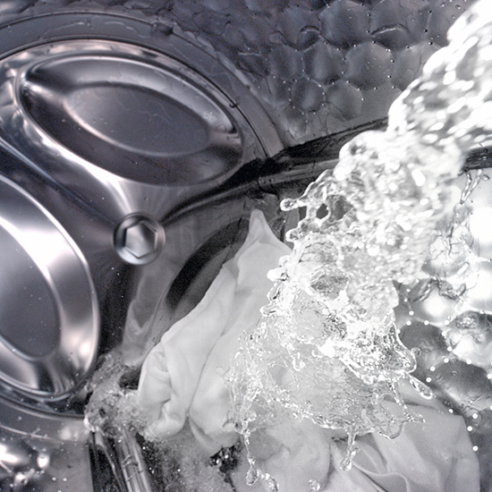 Im Vordergrund ist eine Waschmaschine aus Metall mit einem weißen Tuch und Wasserstrahlen zu sehen.