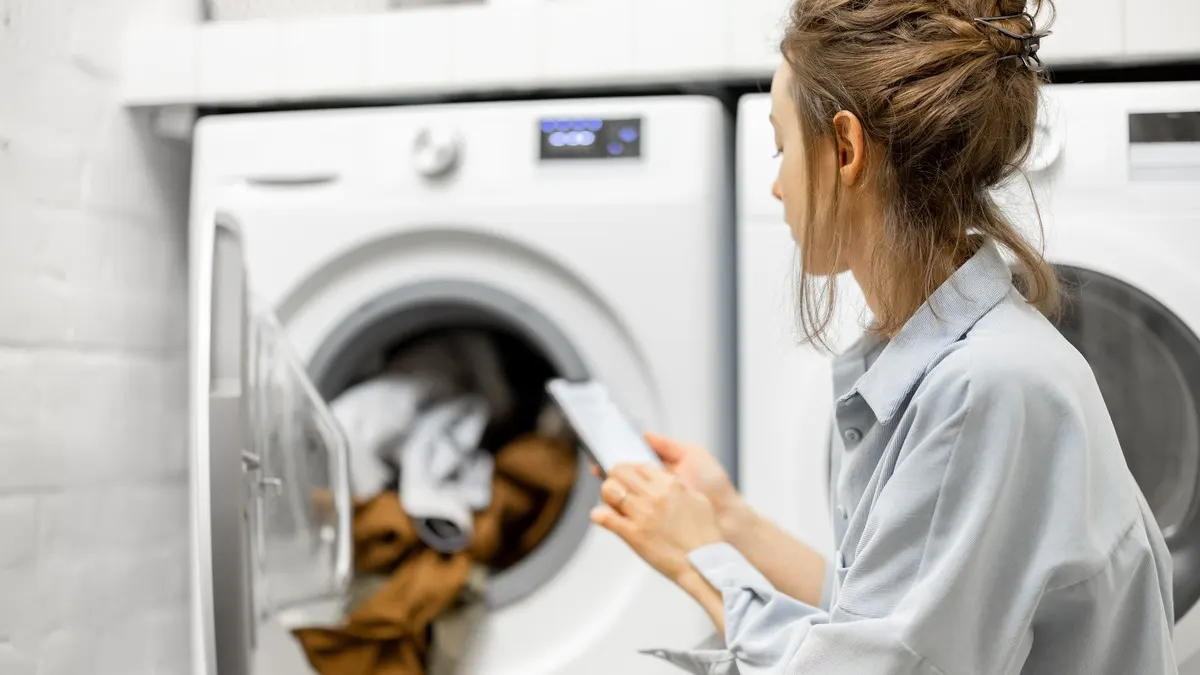 Eine Frau bedient ein Smartphone, während sie vor einer offenen Waschmaschine sitzt.