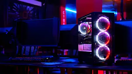 Ein PC-Gehäuse mit drei beleuchteten Lüftern steht auf einem Schreibtisch, neben Monitoren und einer Tastatur in einer dunklen Umgebung.