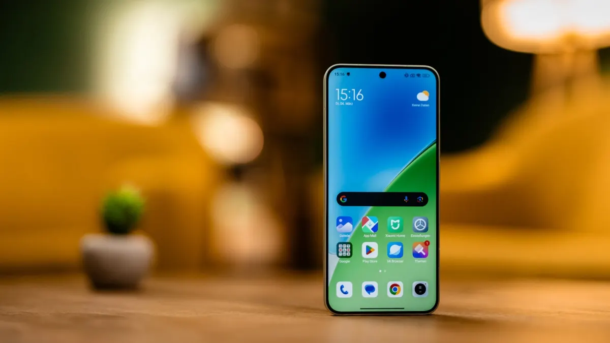 Ein Smartphone mit blau-grünem Bildschirm steht auf einem Tisch; im Hintergrund sind eine Pflanze und eine Lampe zu sehen.