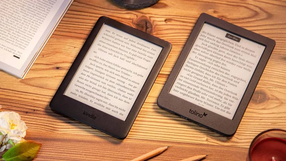Zwei E-Reader, Kindle und Tolino, liegen auf einem Holztisch, mit Text auf den Bildschirmen. Eine Blume und ein Stift sind auch sichtbar.