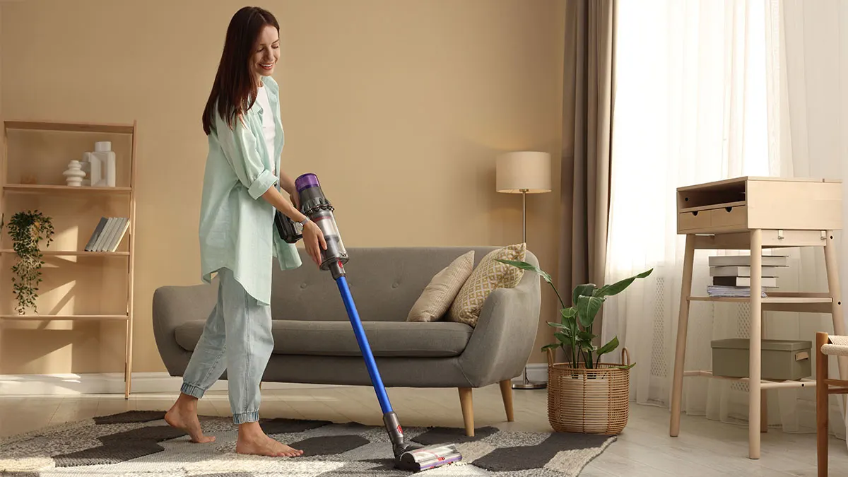 Frau reinigt mit einem blauen Stabstaubsauger den Teppich vor einem Sofa im Wohnzimmer.