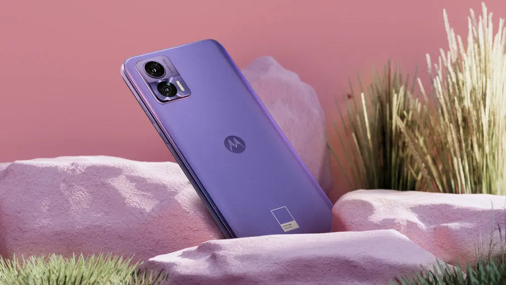 Lila Handy liegt auf rosa Steinen, mit Kameras oben links und einem Logo in der Mitte. Gras im Vorder- und Hintergrund.